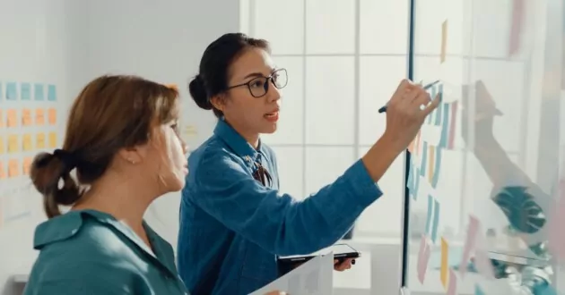 مجموعة من سيدات الأعمال الآسيويات الشابات يرتدين ملابس غير رسمية أنيقة باستخدام الكمبيوتر اللوحي ويكتبن على ملاحظات لاصقة ملونة لتطوير مشروع تجاري في مكتب إبداعي.