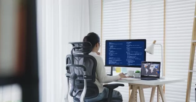 Жінка сидить за столом, спиною повернувшись до камери, працює на настільному комп’ютері