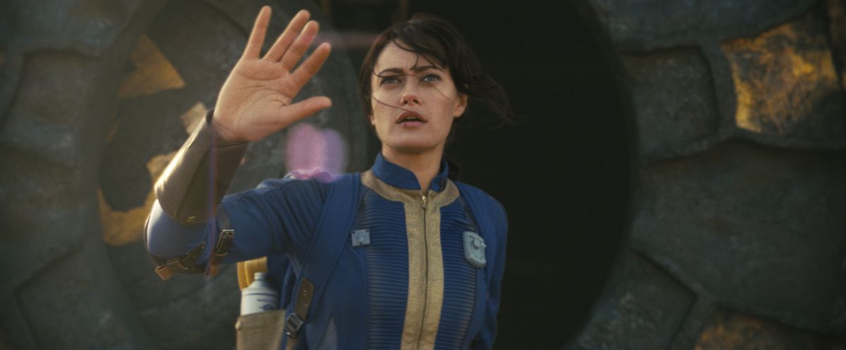 Lucy (Ella Purnell) holding her hand up and looking at something as she emerges from Vault 33