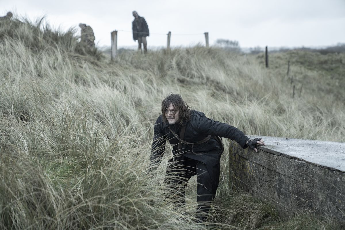 Daryl (Norman Reedus) crouching in some tall grass with zombies looming in the background behind him in a still from The Walking Dead: Daryl Dixon