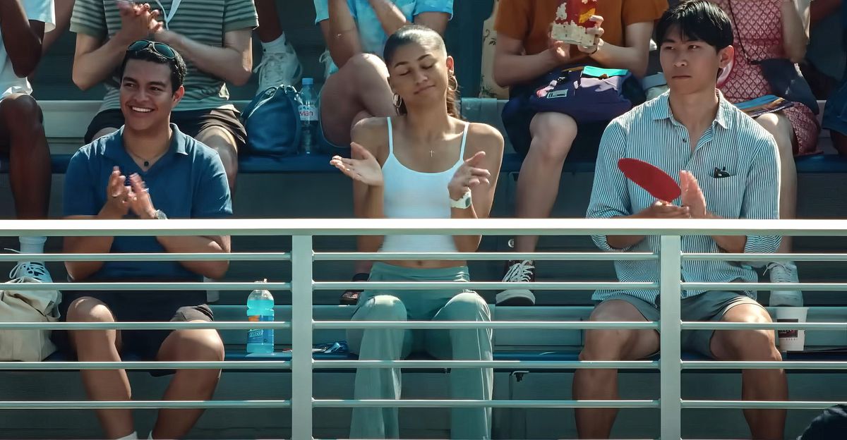Tennis player Tashi (Zendaya) sits in the stands at a match in Luca Guadagnino’s Challengers. The fans around her are applauding something going on on the court, but she’s smiling and shrugging, with her eyes closed.