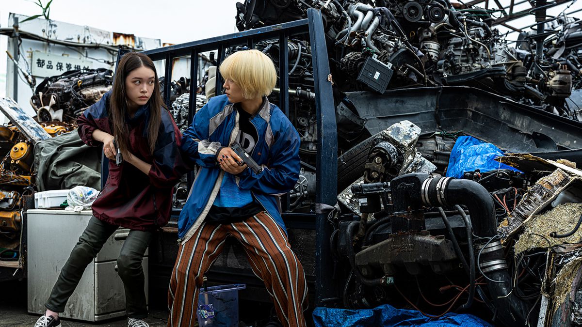Akari Takaishi and Saori Izawa hold pistols and take cover behind a pile of trashed car parts in Baby Assassins 2
