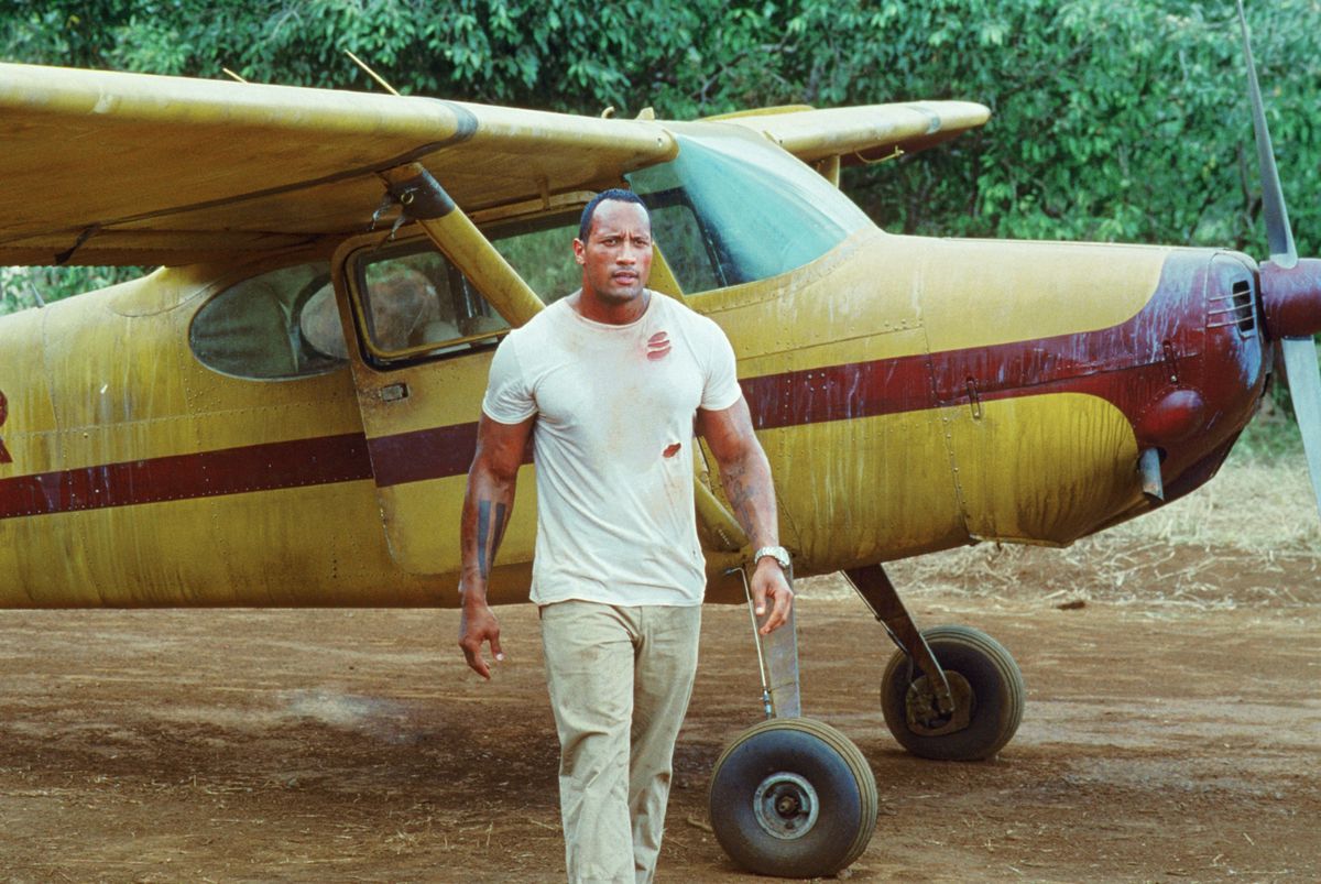 Dwayne “The Rock” Johnson stands in a dirty T-shirt and khakis in front of a small bush plane in the film The Rundown.