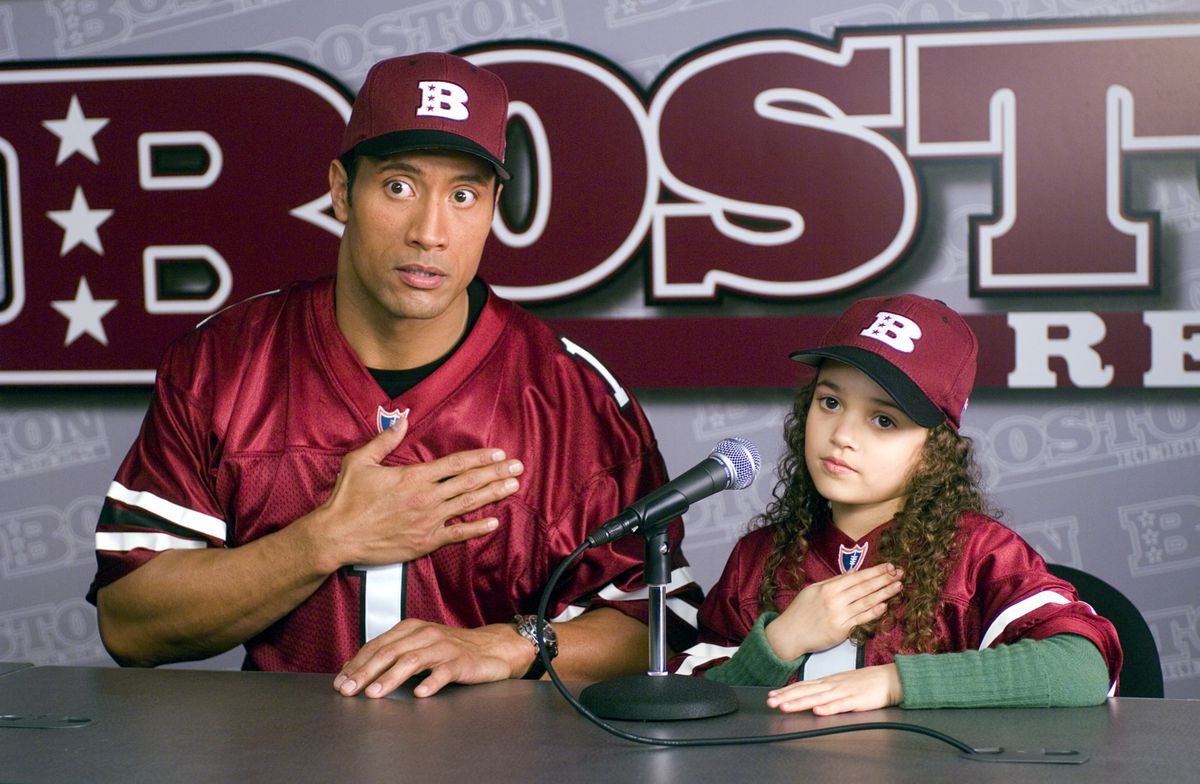 Dwayne Johnson plays a football player at a press conference next to his daughter, in a matching jersey and cap, in the film The Game Plan