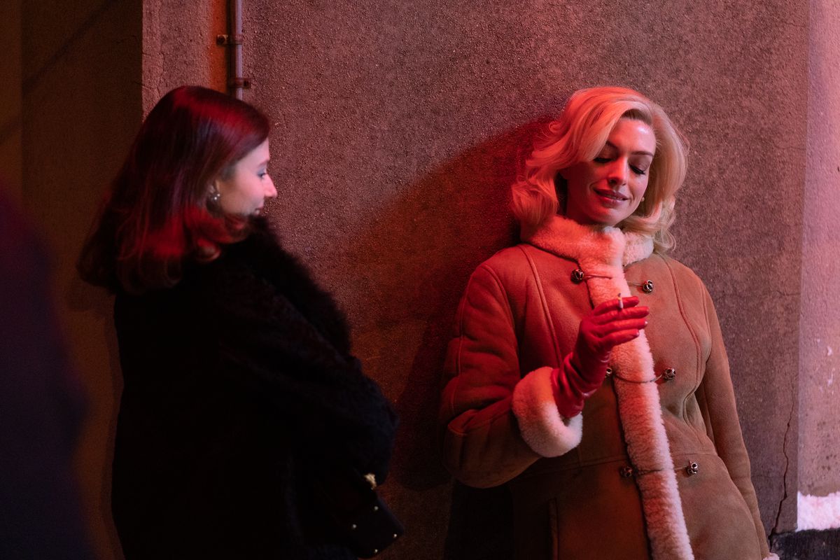 Anne Hathaway, in a blond wig and shearling coat, smokes leaning against a neon-drenched wall as Rebecca while Thomasin McKenzie looks on in the movie Eileen.
