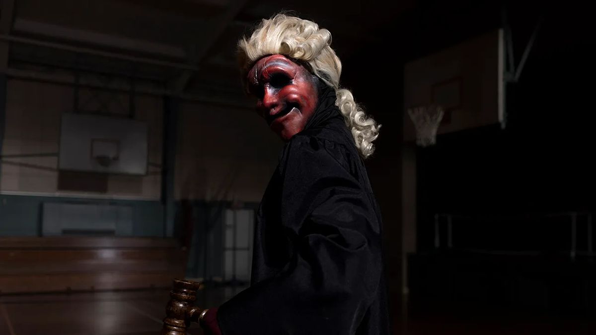A masked figure in a black cloak and white wig holding a gavel in a dark gymnasium in Founders Day.