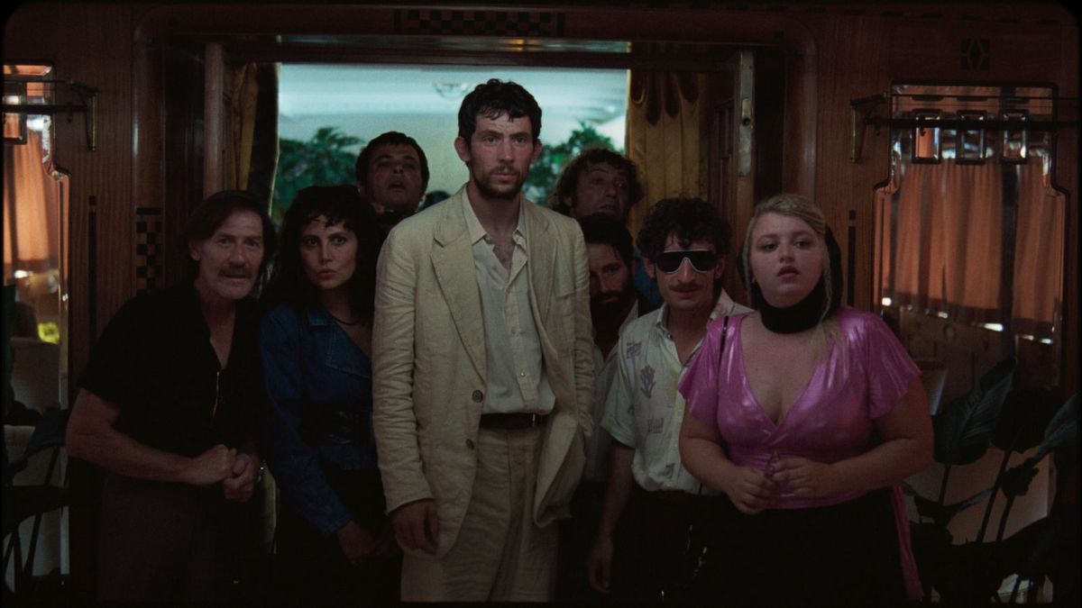 A man in a white, wrinkled suit with an open collar button shirt surrounded by a group of people looking at something off-screen with fascination.