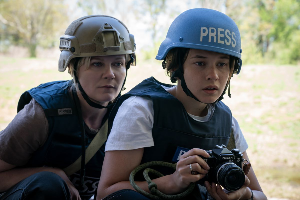 Photojournalists Lee Miller (Kirsten Dunst) and Jessie (Cailee Spaeny) huddle together, Jessie clutching her camera and wearing a bright blue helmet that says “PRESS,” in Alex Garland’s Civil War