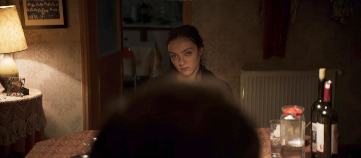 A woman sitting at a dinner table, dimly lit. She looks vaguely pissed off. We are looking at her over someone else’s head.