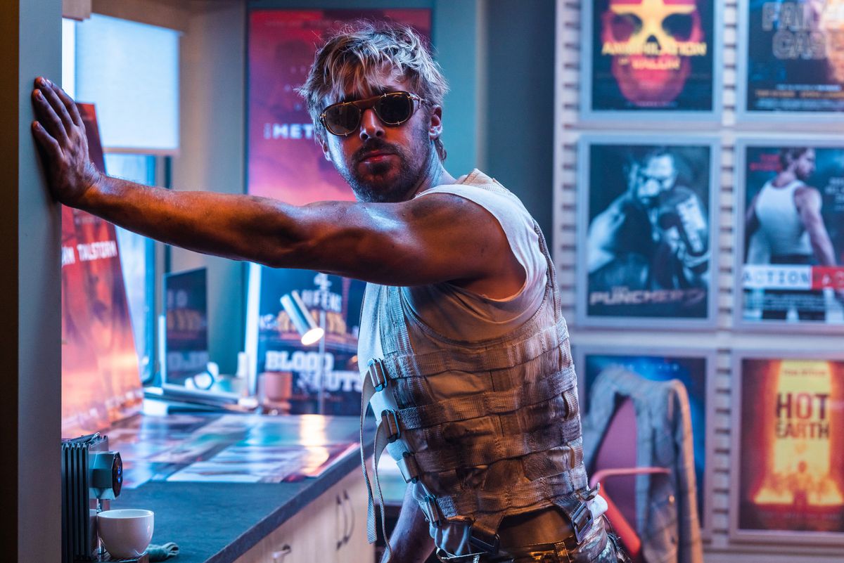 In a scene from The Fall Guy, stunt man Colt Seavers (Ryan Gosling, in sunglasses, a filthy sleeveless vest, and a scowl) leans one-armed against a wall in a room lined with neon-colored movie posters