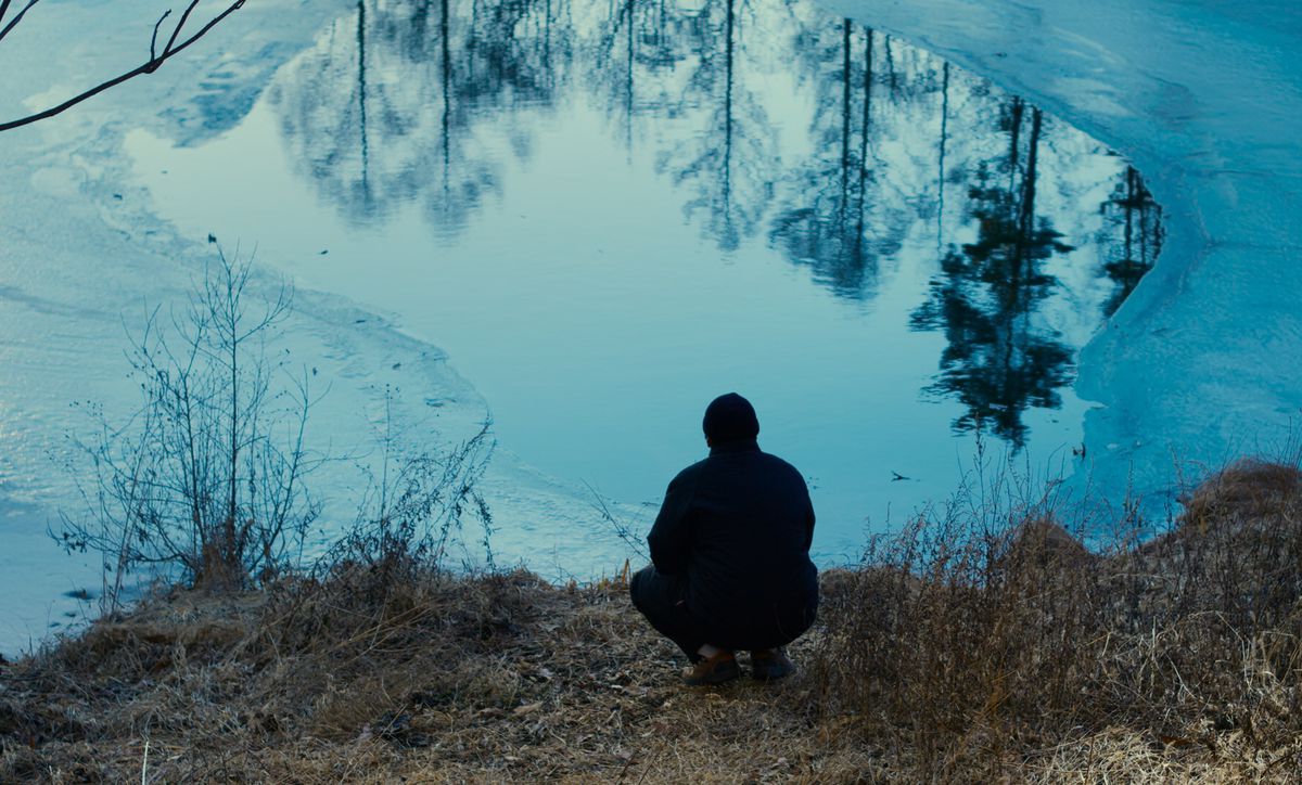 A figure in a coat and beanie seen crouching from behind at the edge of a half-frozen lake, the woods reflecting on its surface in the film Evil Does Not Exist.