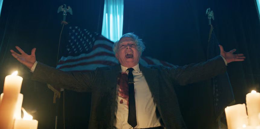 Brad Dourif, with blood on his shirt, screams with glee as the American flag waves behind him in Chucky
