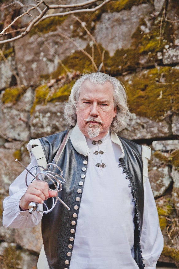 A white haired man with a pointy goatee standing in front of a stone while and dressed like a musketeer, as if pulled from the pages of Alexandre Dumas' novels. He's in a white shirt with a leather waistcoat of sorts, and he has a resting on his shoulder. It is a strong look.