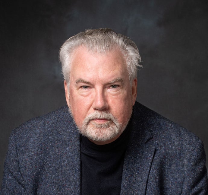 A posh headshot photograph of Lawrence Schick. He's a white man with brushed back white hair an a neat white goatee. He's wearing a black top of some kind under a speckled dark blue blazer. He's leaning forward slightly, as if looking into the viewer's soul.