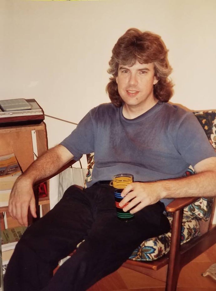 A photo of a bushy haired - possibly mulleted - man slouched in a cushioned, wooden chair with a cup of some kind of drink in his hand, looking at the camera. It looks so faded it could be an image from the 80s, but it's from the mid 90s. It's Lawrence Schick.