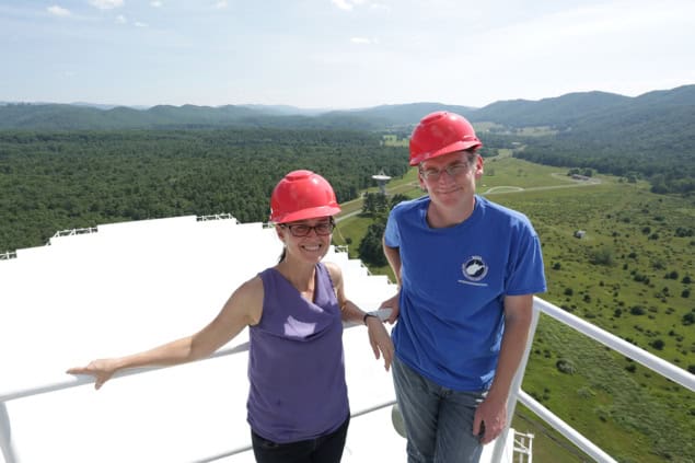 Two people wearing red hard hats stood on a platform overlooking a telescope