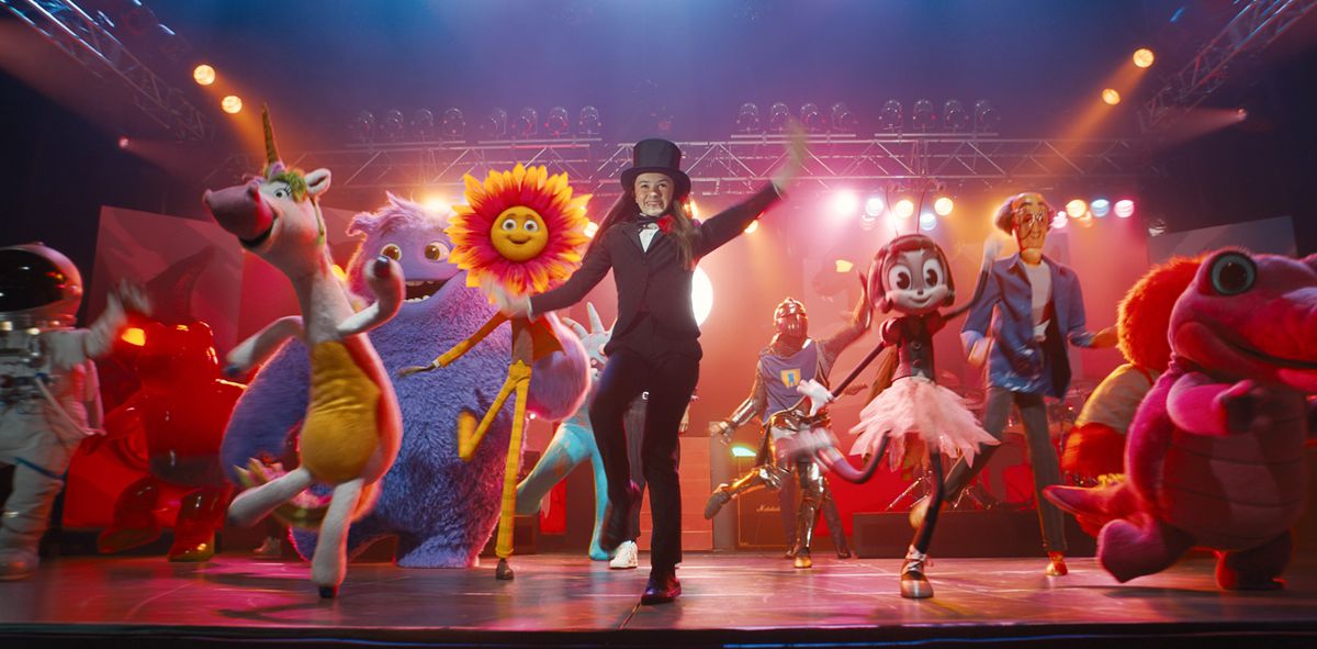 Bea (Cailey Fleming), in suit and top hat, dances on a stage under bright lights, surrounded by CG creations representing kids’ imaginary friends: a big-nosed bipedal unicorn in a yellow body suit, an astronaut, a sunflower with a face and legs, a big purple furry creature, and more