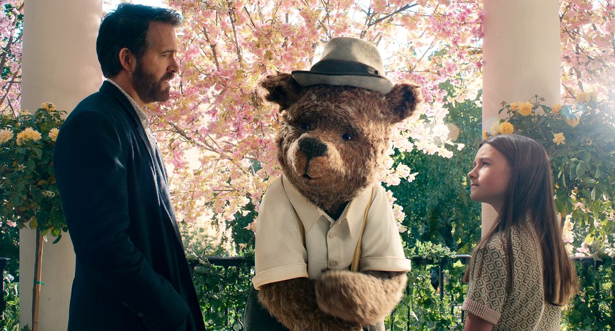 Cal (Ryan Reynolds) and Bea (Cailey Fleming) stand outdoors, surrounded by flowering trees and bushes, both facing a human-sized CG imaginary-friend character who looks like a teddy bear in a battered fedora and button-up shirt in John Krasinski’s IF