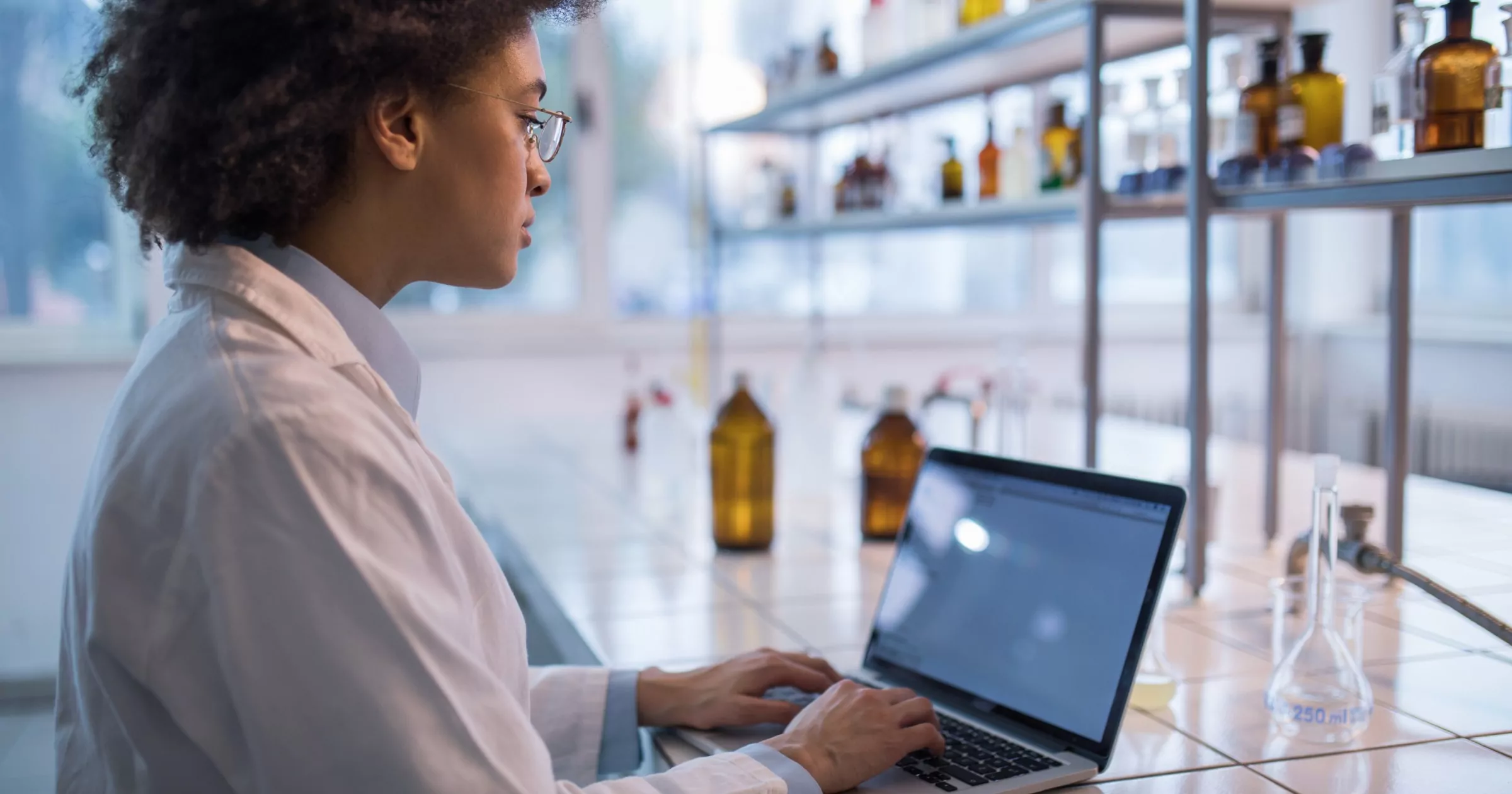 Doctor working on laptop with medicine bottles and beakers on shelves