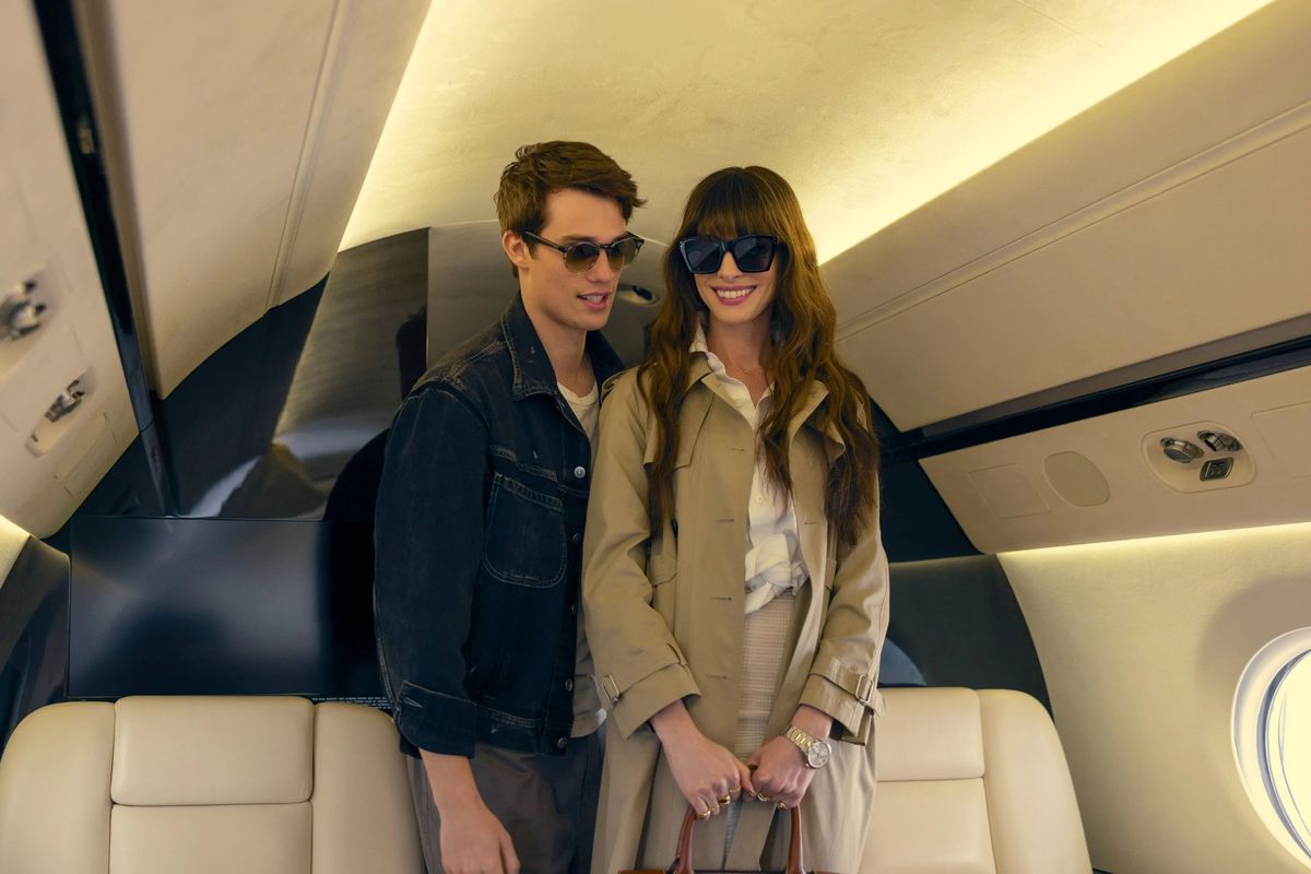 A man in a jean jacket wearing sunglasses stands next to a smiling woman in a tan long-sleeve jacket with sunglasses on in The Idea of You.