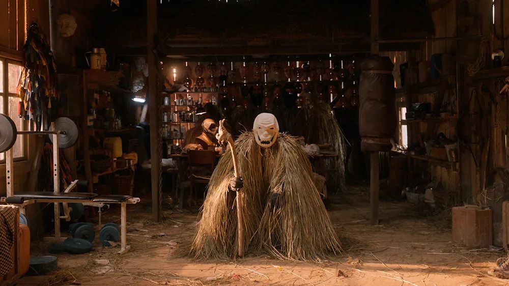 A person wearing a leering mask and straw costume holding a staff in the middle of a workshop in Pamfir.