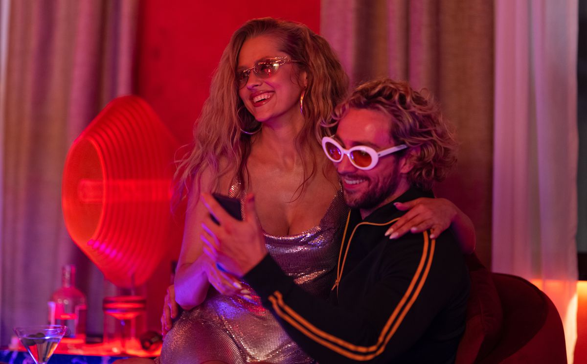 In a scene from The Fall Guy, movie star Tom Ryder (Aaron Taylor-Johnson, in black track suit and white plastic sunglasses) sits grinning in a red-lit room with a woman in a low-cut silver lamé dress on his lap. But he’s looking at his phone, not her, because he’s a jerk. 