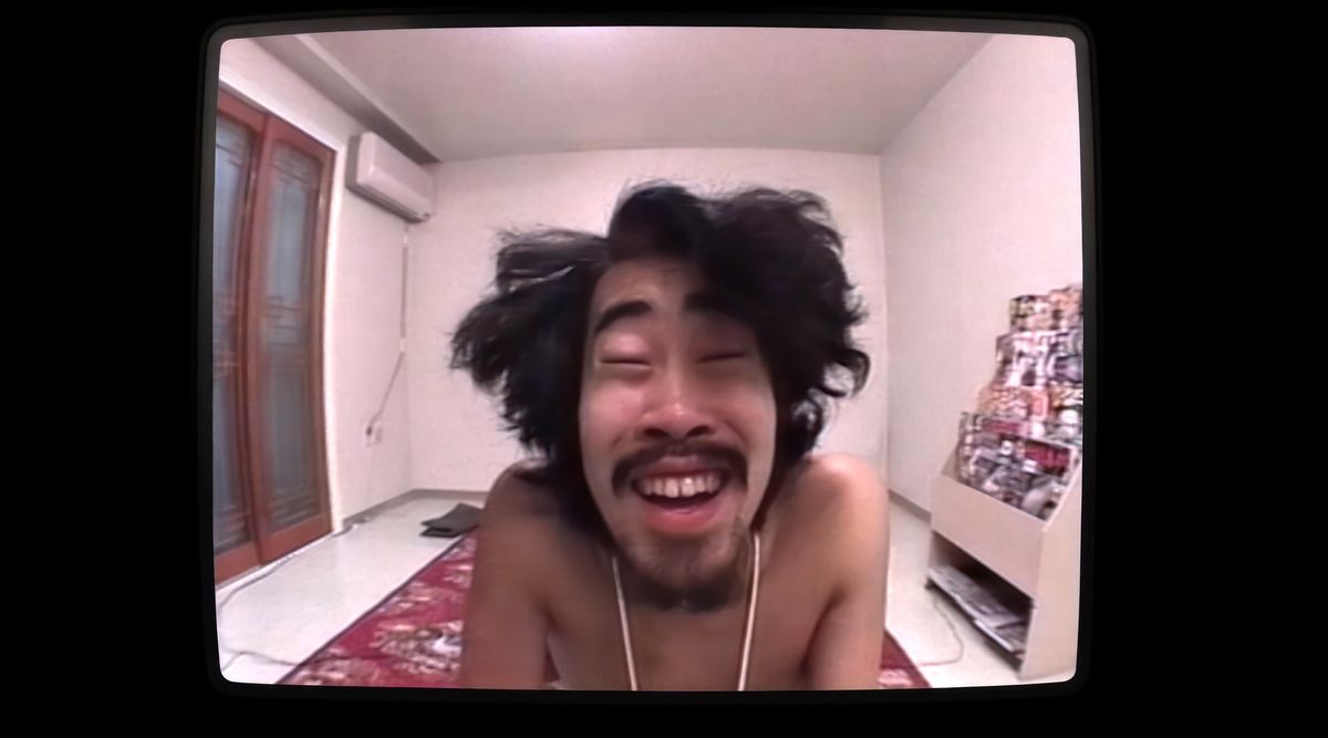 Nasubi, a Japanese man with wild, unkempt long hair, grins into the camera in a scene from Hulu’s documentary The Contestant
