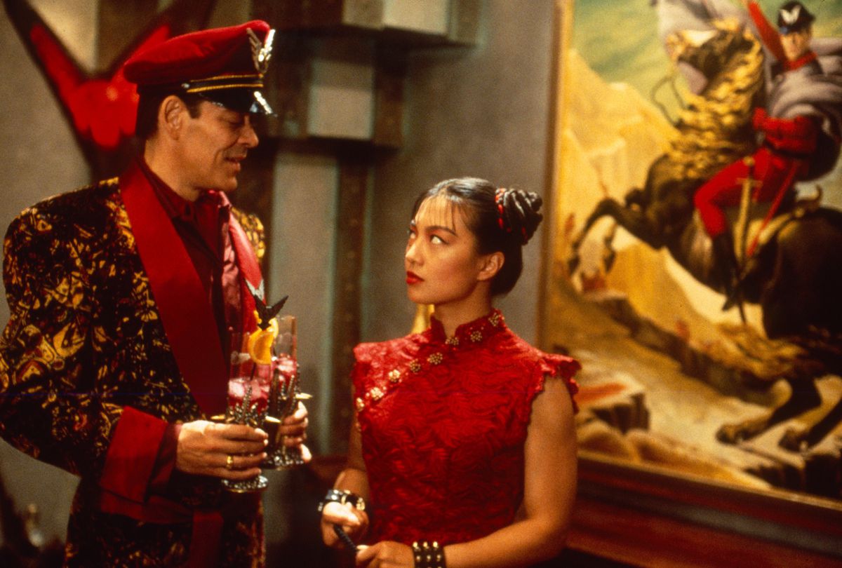 Raul Julia, wearing ridiculous casual villain velour, holds drinks in front of Ming-Na Wen’s Chun-Li, in front of a painting of Raul Julia’s character on a horse, in Street Fighter