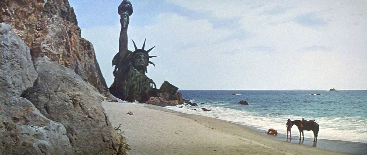 A man slumped over on the shore of beach with a woman and a horse behind him and the ruins of the Statue of Liberty in the background.