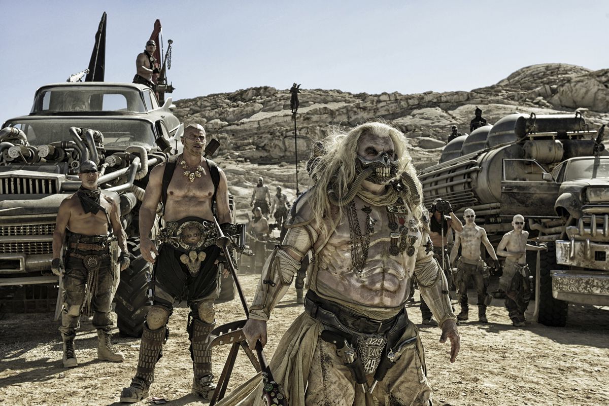 l-r: Nathan Jones as Rictus Erectus and Hugh Keays-Byrne as Immortan Joe stand surrounded by War Boys and huge vehicles in Mad Max: Fury Road.