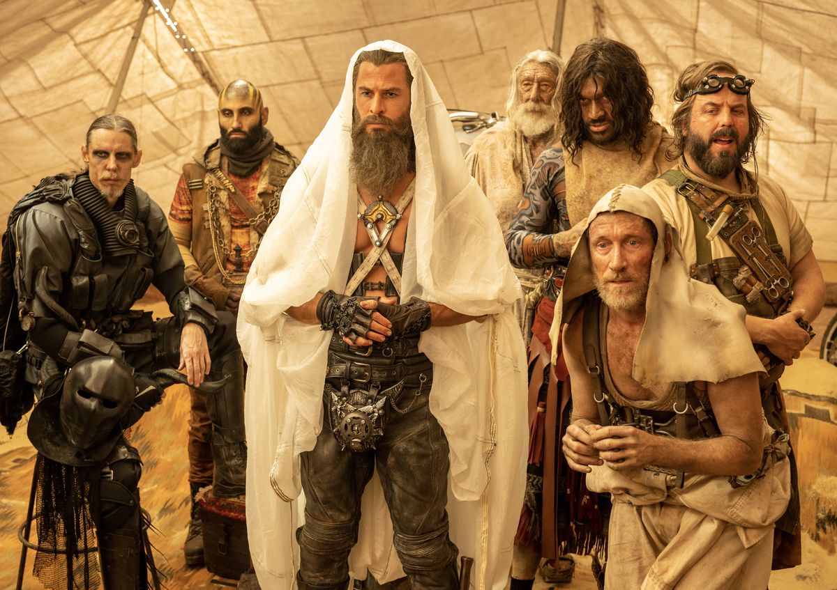 Dementus (Chris Hemsworth), the bearded leader of a group of Wasteland warriors, stands in a tent with a clean white cloth draped over his head and extending down to his feet, surrounded by male acolytes of various ages in George Miller’s Furiosa