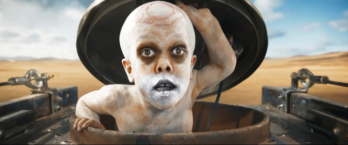 War Pup (Quaden Bayles, shirtless, painted white, with chrome spray paint across his mouth and circular scarification on his skull) pops out of a hatch in the War Rig in Furiosa