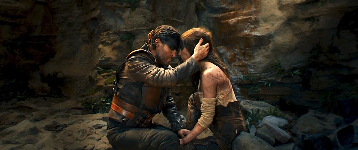 Praetorian Jack (Tom Burke) and Furiosa (Anya Taylor-Joy) sit close to each other in a barren, rocky cave, holding hands and with their heads pressed together, in Furiosa